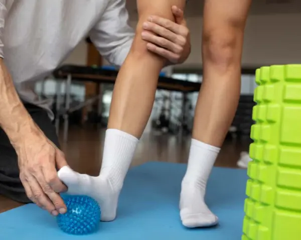 Fizjoterapeuta pomaga w rehabilitacji stopy, masując ją kolczastą piłeczką. Obok zielony wałek do ćwiczeń.