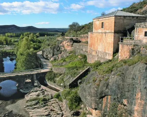 El portón de la oliva, un antiguo edificio de piedra junto a un río y un puente de arco, se alza sobre un paisaje rocoso y verde.