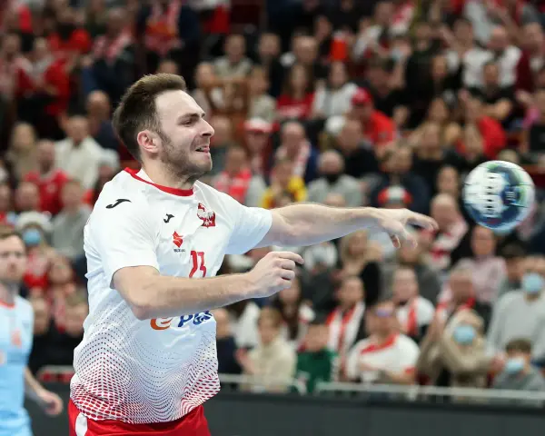 Piłkarz ręczny Polski w akcji, rzuca piłkę. W tle widać trybuny z kibicami, którzy dopingują swoją drużynę w meczu przeciwko Iranowi.