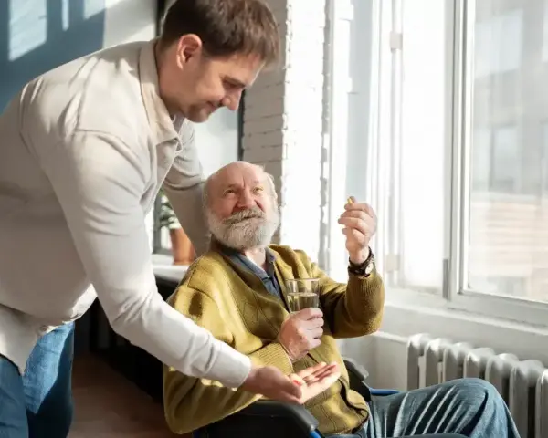 Mężczyzna w średnim wieku podaje starszemu mężczyźnie w wózku inwalidzkim tabletkę i szklankę wody.