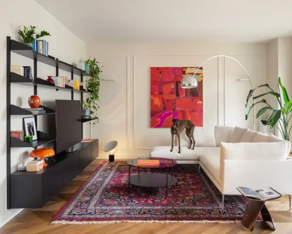 Un galgo se posa sobre un sofá blanco, rodeado de arte abstracto y un tapete persa. El **ruido visual** de los objetos decorativos y la pintura vibrante crea un ambiente ecléctico.