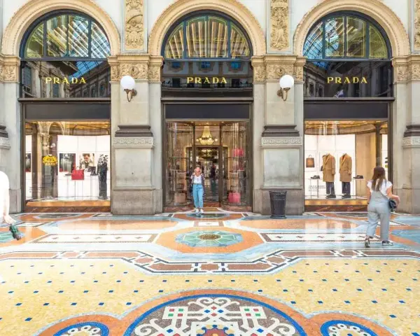 Ingresso del negozio **Prada** con vetrine eleganti e un mosaico colorato sul pavimento. Persone passeggiano davanti alla boutique.