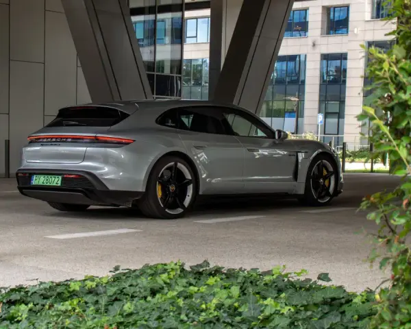 Szare Porsche Taycan Turbo S stoi pod zadaszeniem, z widokiem na nowoczesny budynek i zieleń.