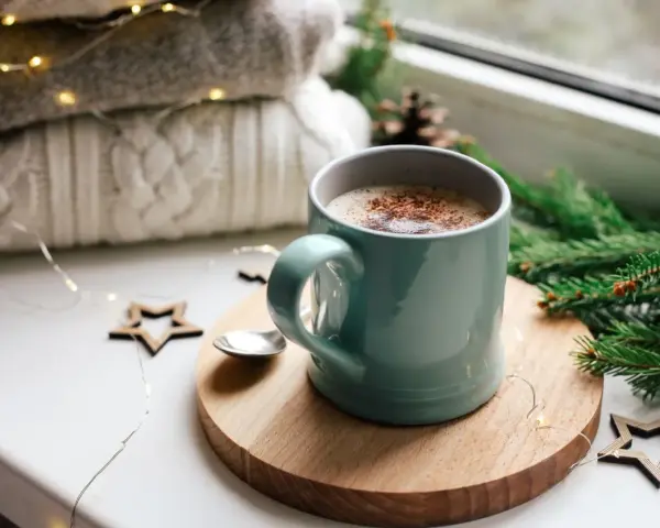Une tasse de chocolat chaud fumant, entourée de décorations de Noël et de lumières scintillantes, promet un moment de réconfort.
