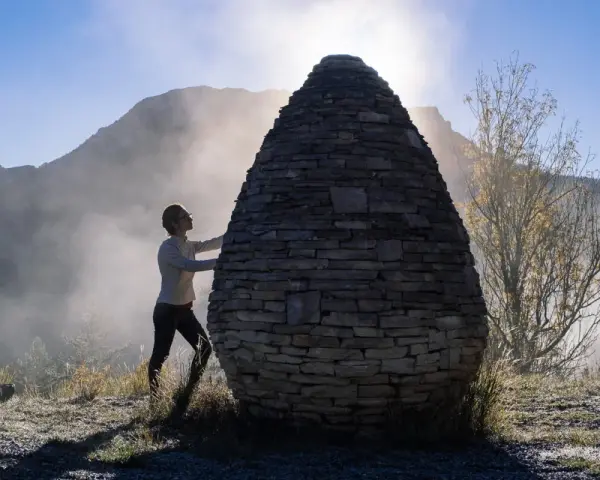 Une femme interagit avec une structure ovoïde en pierres, une œuvre de land art, dans un paysage brumeux et montagneux au lever du soleil.