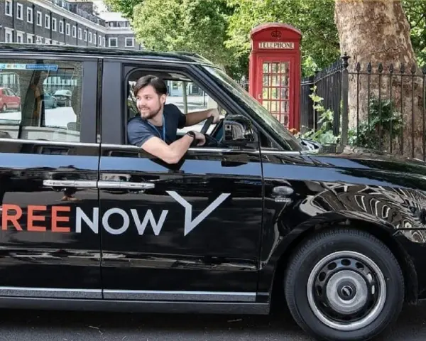 Ein Mann fährt ein schwarzes freenow taxi. Im Hintergrund steht eine rote Telefonzelle.