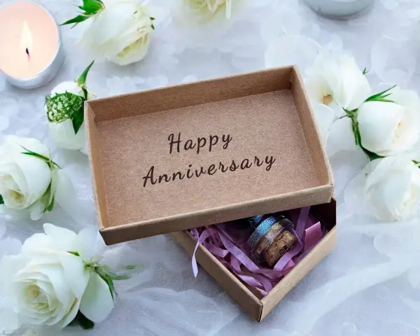 Otwarta szkatułka z napisem "Happy Anniversary" i małą buteleczką w środku, otoczona białymi różami i płonącą świecą. Idealny prezent na 21 rocznicę ślubu.