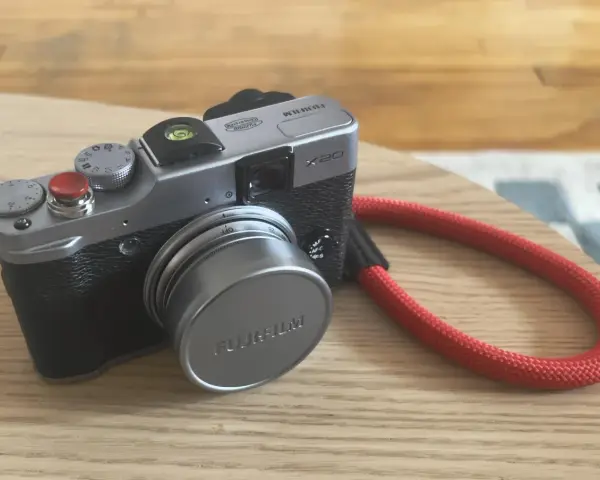Srebrny aparat Fujifilm X20 z czerwoną smyczą leży na drewnianym blacie.
