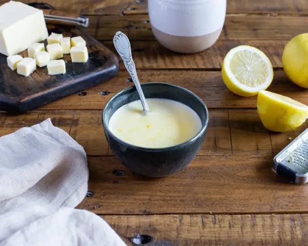 Un bol de sauce beurre citron, avec des citrons, du beurre coupé en dés et une râpe à zester sur une table en bois.