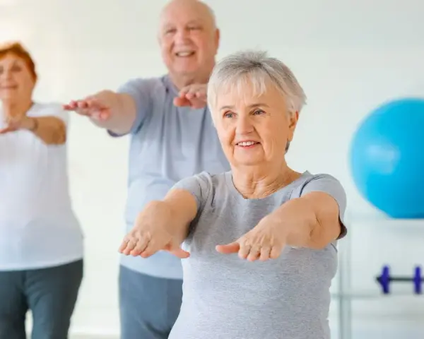 Trois seniors souriants pratiquent la gym douce, bras tendus, dans une salle lumineuse avec un ballon bleu en arrière-plan.