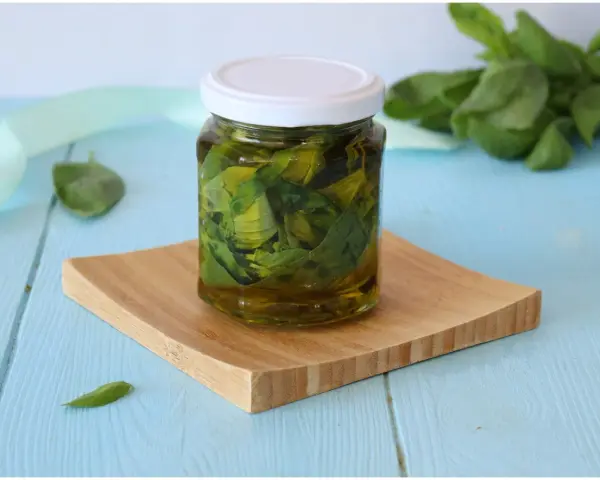 Un barattolo di olio al basilico fatto in casa, con foglie fresche immerse nell'olio dorato, su un tagliere di legno.