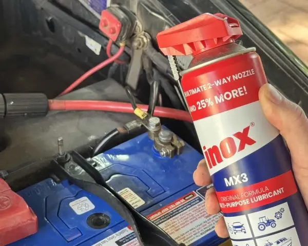 A hand holds a can of INOX MX3 lubricant near a car battery. Does cleaning battery terminals help? This spray might be the answer.