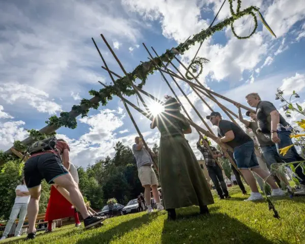 Midsommarstången reses av en grupp människor, en traditionell del av hur firar man midsommar. Solen skiner genom molnen.