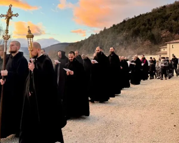Monks in black robes process with a cross and lanterns, their solemn religious chants echoing as they walk towards a building at sunset.