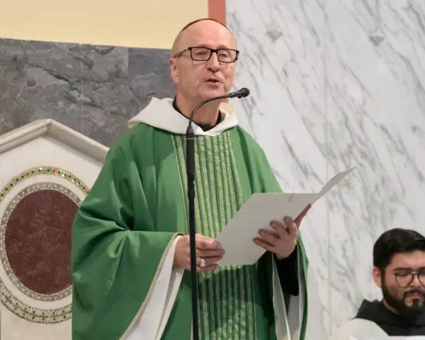 A man with a Benedictine title, wearing a green chasuble, speaks into a microphone while holding a paper.