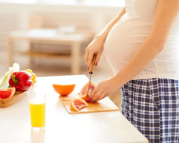 Schwanger, die Grapefruit für einen gesunden Snack schneidet. Nebenan liegen Paprika und ein Glas Saft.