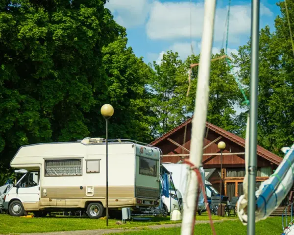 Słoneczny dzień na kempingu na Mazurach. Kamper stoi na trawie obok drewnianego budynku, w tle zielone drzewa i plac zabaw.