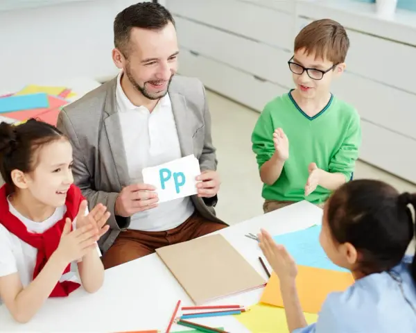 Nauczyciel pokazuje literę "Pp" podczas zebrania z rodzicami. Dzieci klaszczą, ucząc się alfabetu.