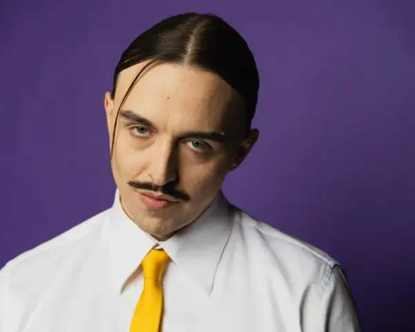 Tommy Cash w białej koszuli i żółtym krawacie, z charakterystycznym wąsem i przedziałkiem na środku głowy, na fioletowym tle.