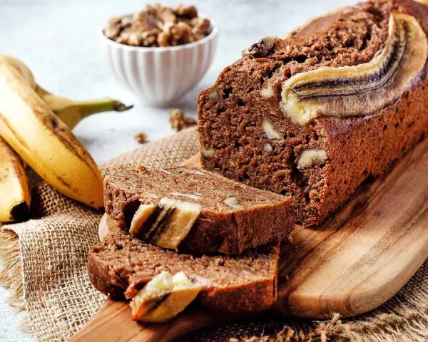 Saftiges, veganes Bananenbrot mit Walnüssen und Bananenscheiben, daneben frische Bananen und eine Schüssel Nüsse.