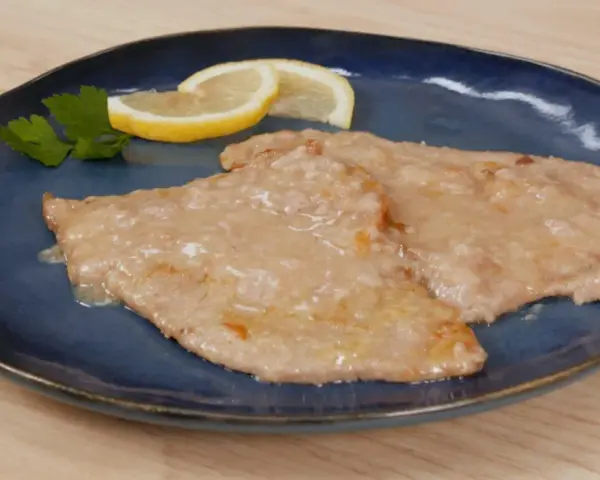 Due scaloppine al limone cremose, guarnite con prezzemolo e spicchi di limone, su un piatto blu.