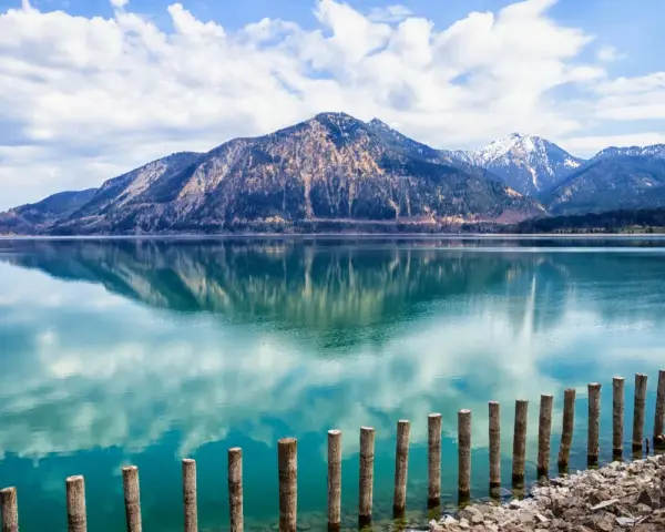 Spiegelglatter See mit Bergen im Hintergrund, wie man ihn in Bayern sehen kann. Holzpfähle ragen aus dem türkisfarbenen Wasser.