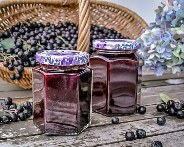 Zwei Gläser mit Aronia-Marmelade, bereit zum Verarbeiten. Ein Korb voller Aronia-Beeren steht im Hintergrund.