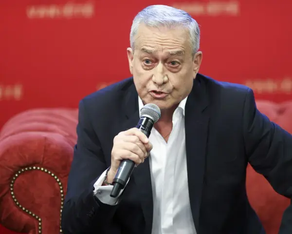 Janusz Szydłowski przemawia do mikrofonu, siedząc na czerwonej, pikowanej kanapie.