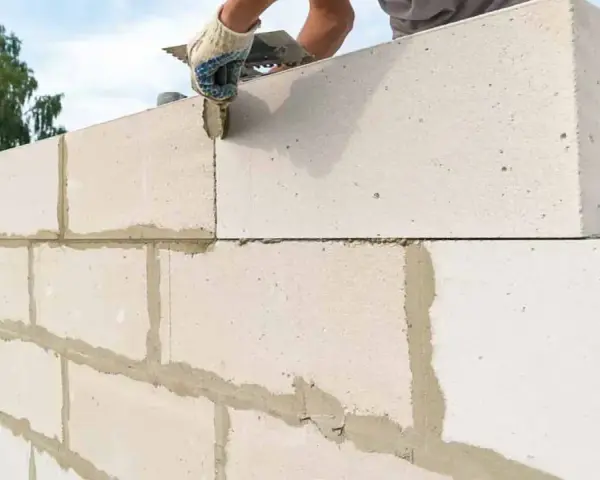 Budowanie ściany z bloczków betonowych. Ręka w rękawicy nakłada zaprawę na kolejny element.
