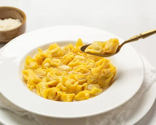 Un cucchiaio d'oro solleva un boccone di cappelletti in brodo fumante da un piatto bianco.