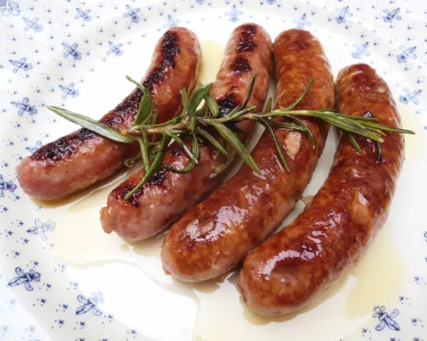 Salchichas al vino, doradas y jugosas, reposan sobre un plato blanco con delicados motivos azules, adornadas con ramitas de romero fresco.