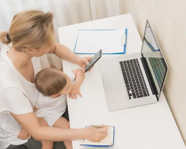 Kobieta z dzieckiem na kolanach sprawdza na telefonie informacje o zasiłku macierzyńskim 80 czy 100% i 60. Obok laptop i notatnik.