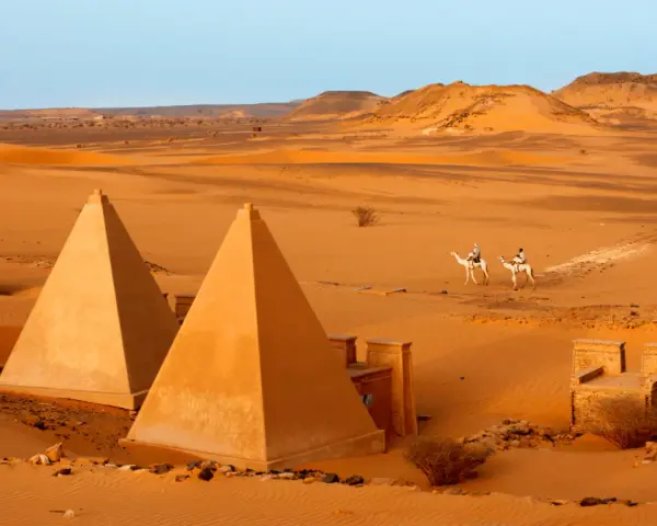 Pustynny krajobraz Sudanu z piramidami i dwoma podróżnikami na wielbłądach.