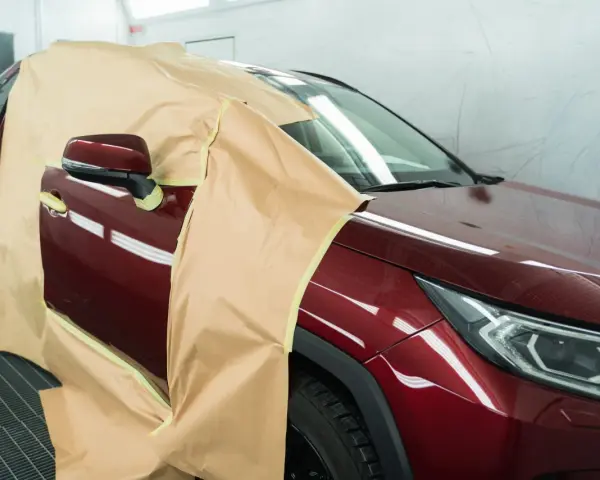 Voiture rouge bordeaux masquée pour la peinture. On se demande combien de temps entre peinture et vernis pour un tel travail.