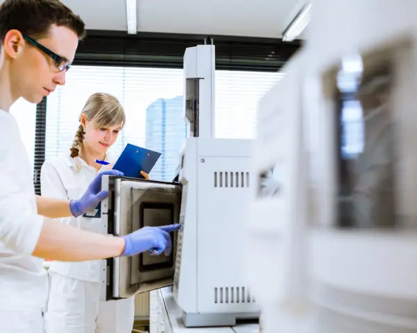 W laboratorium młoda para naukowców pracuje przy aparaturze. On w okularach, ona z notatkami.