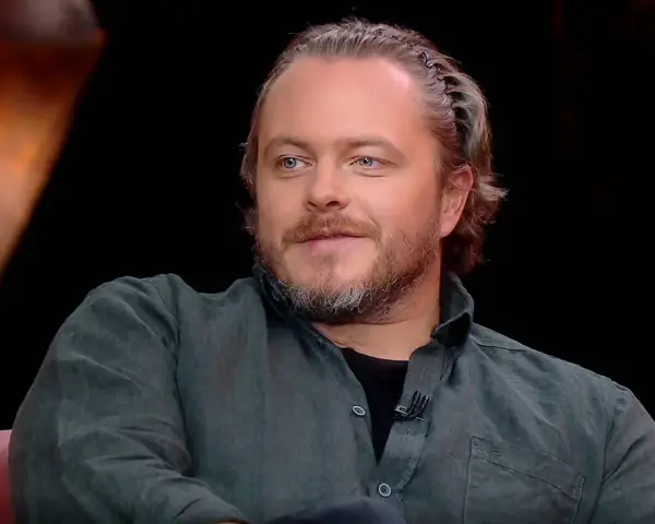 Tomasz Schuchardt, znany z wielu filmów, seriali i programów, z charakterystyczną fryzurą i niebieskimi oczami, siedzi na kanapie.