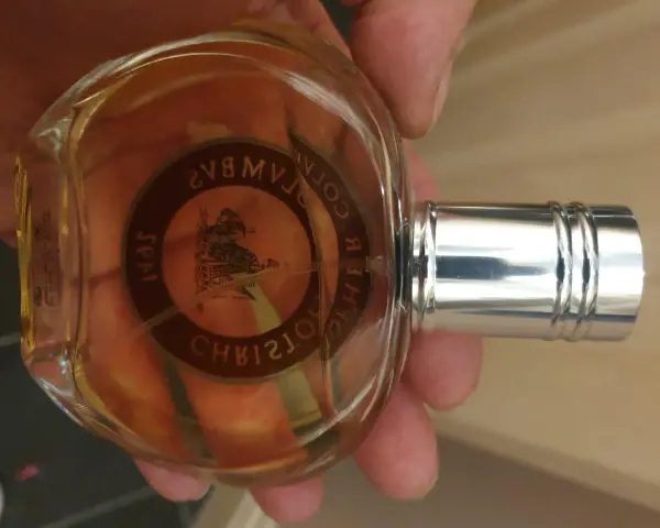 Szklana butelka z bursztynowym płynem, ozdobiona okrągłą etykietą z wizerunkiem statku i napisem "Christopher Columbus perfumy".