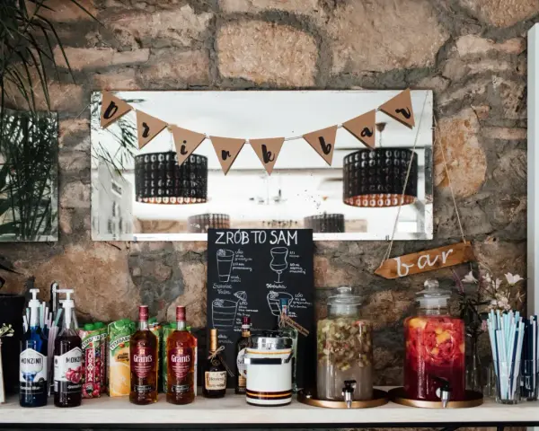 Stół z napojami i składnikami do drinków, tworzący domowy co zamiast drink baru. Na półce stoją butelki alkoholu, syropy, kartony z sokami i dwa duże słodkie poncze.