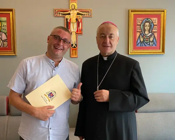 Uśmiechnięty mężczyzna z dokumentem i biskup pozują na tle krzyża i obrazów. Mężczyzna trzyma dokument z herbem, a biskup ma na sobie sutannę.