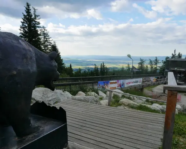 Pomnik niedźwiedzia na tle panoramy Tatr poza szlakiem. Drewniana kładka prowadzi do budynku z widokiem na dolinę.