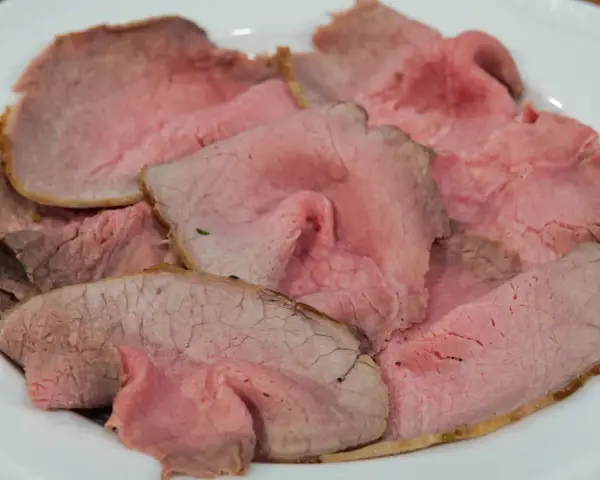 Fette sottili di arrosto di manzo rosa e succulento, servite su un piatto bianco.