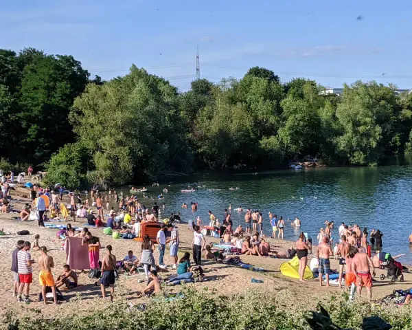 Ein sonniger Tag am **Badesee Bonn**. Viele Menschen genießen das Wasser und den Strand, umgeben von grünen Bäumen.