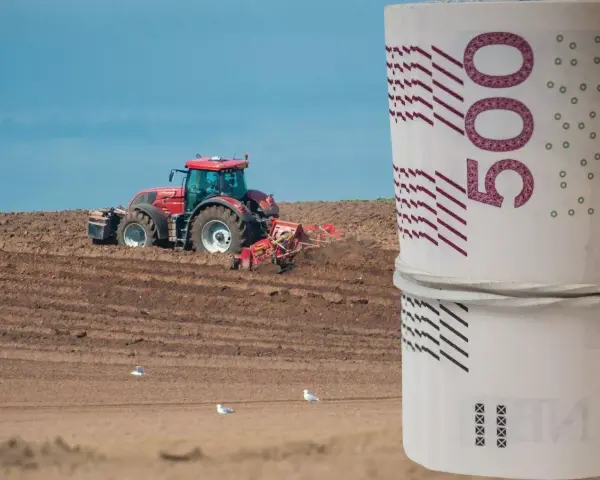 Czerwony traktor orze pole, a na pierwszym planie zwinięte banknoty 500 zł. Zastanawiasz się, ile kosztuje hektar ziemi budowlanej?
