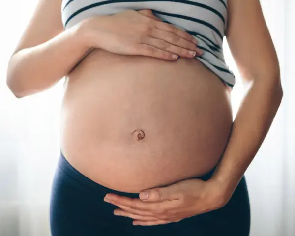 Brzuszek w ciąży z widocznym pępkiem, otoczony dłońmi przyszłej mamy.
