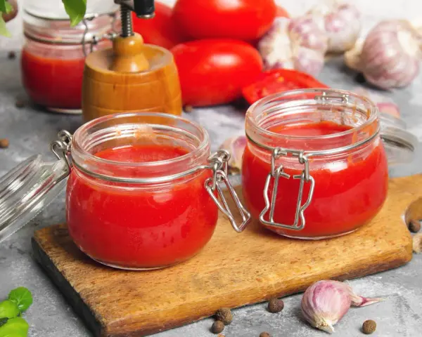 Deux bocaux en verre remplis de coulis de tomates, sur une planche en bois. Des tomates, de l'ail et du basilic complètent la scène.