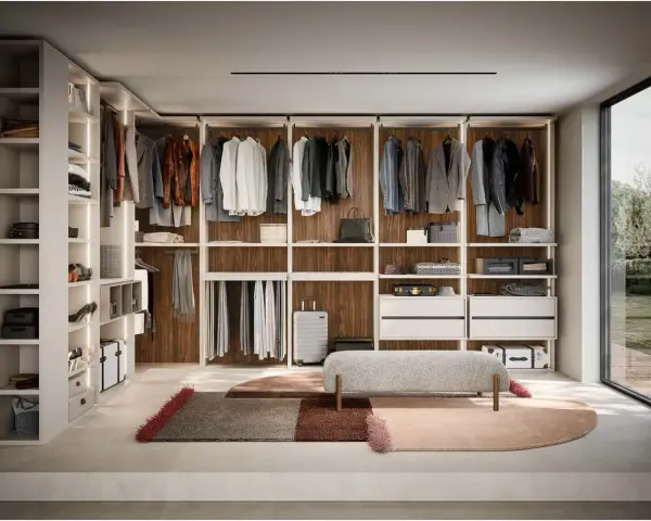 Ampia cabina armadio stanza con scaffali a vista, abiti appesi, cassettiere e un pouf centrale.