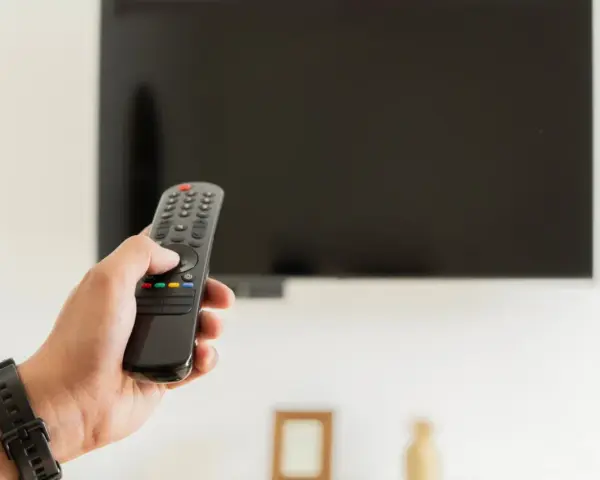 Dłoń trzymająca pilot do telewizora na tle wyłączonego ekranu.