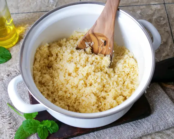 Ein Holzlöffel lockert den frisch zubereiteten Couscous in einem Topf auf. Daneben stehen Öl und Minze.