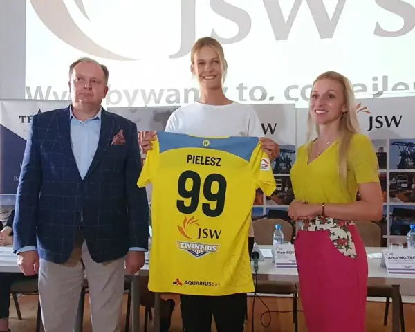 Trzy osoby pozują z koszulką z numerem 99 i nazwiskiem PIELESZ. W tle logo JSW i napisy związane z firmą, być może z okazji wydarzenia w MTS Żory.