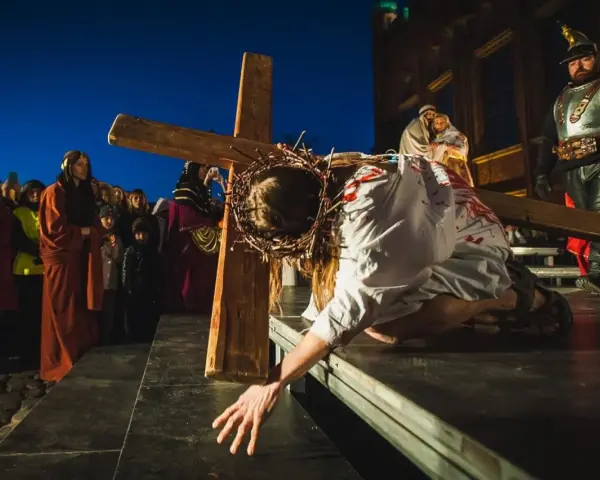 Scena drogi krzyżowej: Jezus w cierniowej koronie, z krwią na szacie, upada pod ciężarem krzyża. Tłum widzów obserwuje to poruszające przedstawienie.
